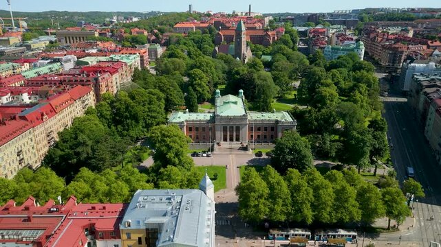 Forward aerial flyover tracking Gothenburg University grand neoclassical facade surrounded by Vasaparken lush canopy with Vasa kyrka and city rooftops visible behind