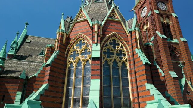 Drone reveals ornate Neo-Gothic facade of Oscar Fredriks Kyrka with intricate tracery windows, gold-framed arches, striped brickwork and verdigris copper details against blue sky