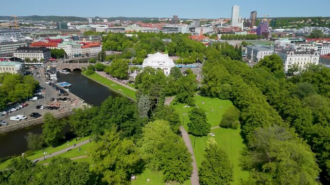 Drone footage flying over lush Kungsparken toward historic Stora Teatern Grand Theater in sunny Gothenburg with canal and city skyline