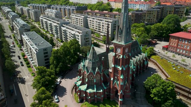 Drone approaches in parallax motion over Oscar Fredriks Kyrka showcasing its ornate Neo-Gothic red brick facade, copper spires and clock tower above Gothenburg cityscape