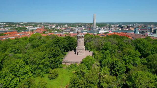 Drone descends toward historic stone Slottsskogen water tower rising above summer tree canopy with sweeping Gothenburg city panorama and harbor visible behind