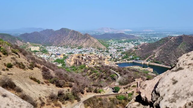 Showing a panoramic aerial view of Amer Fort, Maota Lake, and the surrounding Aravalli hills, with the fort walls winding along the mountains and the city spread across the valley below.