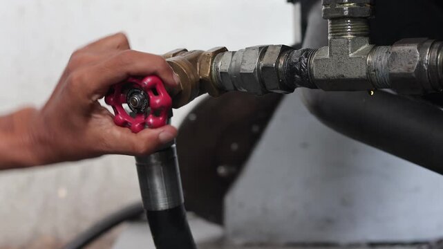 ​A worker's hand turning a red round handle valve on a brass pipe fitting connected to high-pressure hoses in an industrial facility during machine operation.