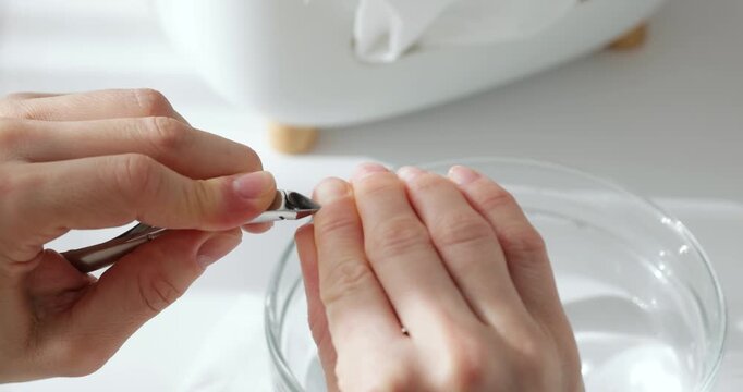 Close up of a woman's hands trimming cuticles with nippers over a bowl of water on white background