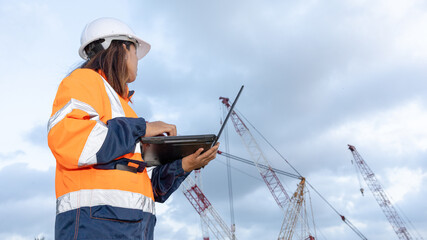 A worker stands at a construction site looking at a laptop while cranes are active in the background. The worker wears safety gear and focuses on the task © Happy Photo