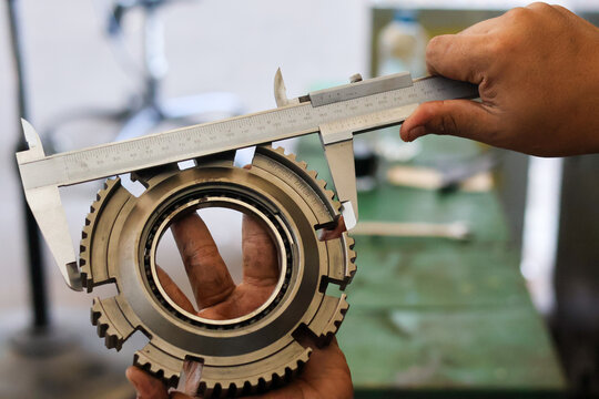 Mechanic hands measuring a steel synchronizer gear using manual vernier caliper in a workshop to check precision dimensions for automotive transmission repair.