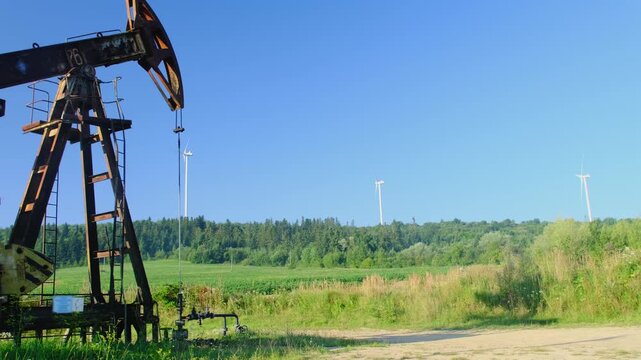Abandoned pumpjack stand in green field against modern wind generators slow motion. Changing fossil fuel with renewable energy for clean life