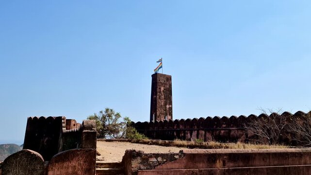 A bright daytime  showing the tall Jaigarh Fort watchtower with a multicolored flag on top, rising above the historic fort walls and open courtyard under a clear blue sky.