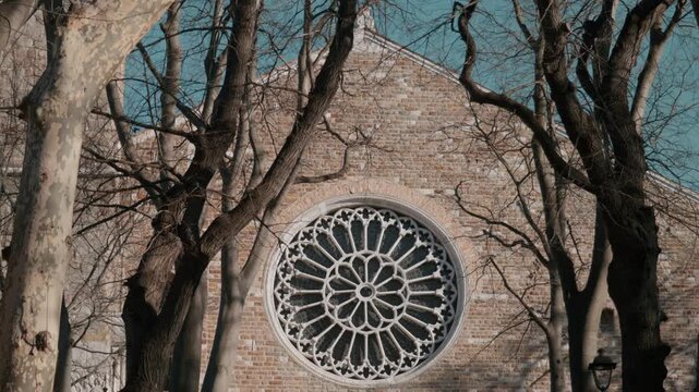 Detailed view of the ancient white stone rose window on the brick facade of the Cathedral of San Giusto in Trieste Italy framed by bare trees on a sunny winter day