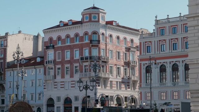 Wide shot of Piazza della Borsa in Trieste Italy with historic palaces fountain statues people walking and vintage street lamps under a bright sunny winter sky