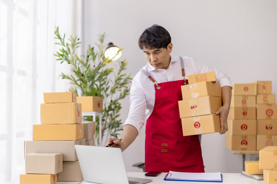 Young Asian man in a red apron running an e-commerce business, checking online orders on a laptop while packing cardboard boxes for shipping and fulfillment from his workspace