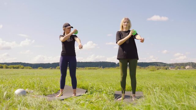 Two young women in sportswear doing myofascial release massage on their forearms and shoulders with a small green ball, standing on yoga mats in a beautiful green field on a sunny day