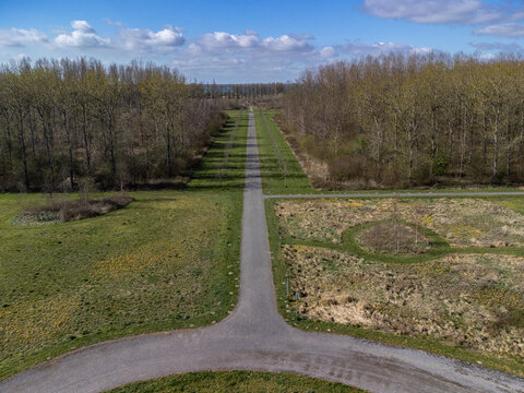 Aerial view of a symmetrical path cutting through green fields and bare trees under a blue sky, De Kiem, Flevoland, Netherlands.