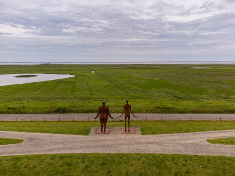 Aerial view of statues standing sentinel over the lush green landscape and distant sea, where the sky meets the horizon, Wachten op hoog water, Friesland, Netherlands.
