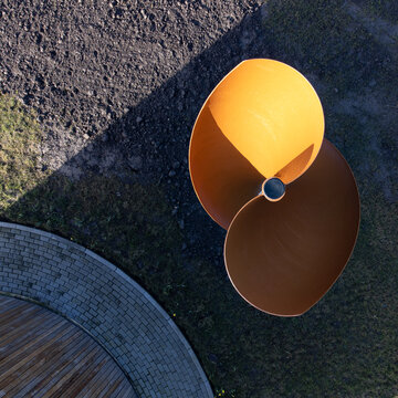 Aerial view of a metallic, orange sculpture casting a long shadow on the green grass near a circular brick structure, Cascadepark, Flevoland, Netherlands.