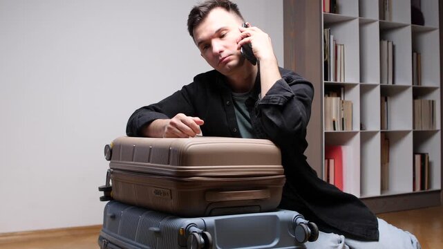 Man sitting on the floor making a phone call, expressing frustration while preparing for travel with stacked suitcases, facing unexpected problems or schedule changes