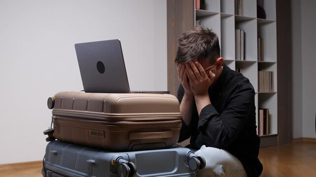 Man covering his face in despair while sitting next to luggage and a laptop, experiencing distress, anxiety, and disappointment over a flight cancellation or disrupted journey