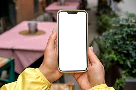 Smartphone mock up held by person wearing  in outdoor cafe setting with tables and greenery in the background, smart phone with white blank screen mobile app design and advertising, online marketing