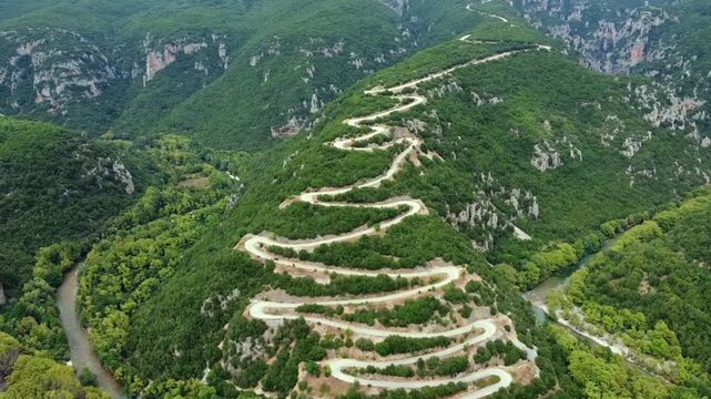 Cinematic drone panorama of the famous hairpin turns in Papigo. High-angle view showing the zig-zag mountain road nestled between the lush banks of the Voidomatis river in Epirus. Ideal for travel.