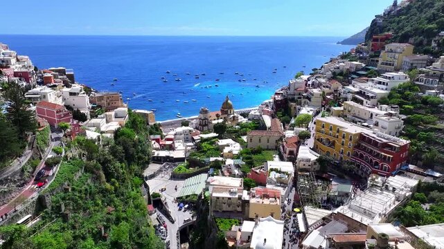 Drone aerial of Positano on Italy's Amalfi Coast, showing cliffside homes, Marina Grande, the Church of Santa Maria Assunta, winding roads, boats, and deep blue Mediterranean water.