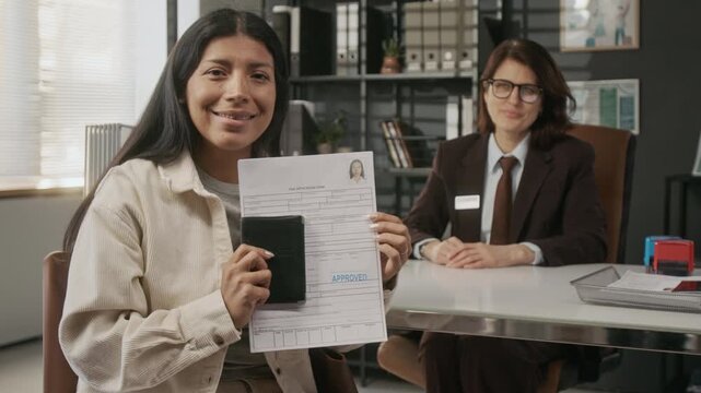 Medium portrait of smiling mature Hispanic woman holding approved visa paperwork after interview with Caucasian female consular officer at embassy, looking at camera with happiness