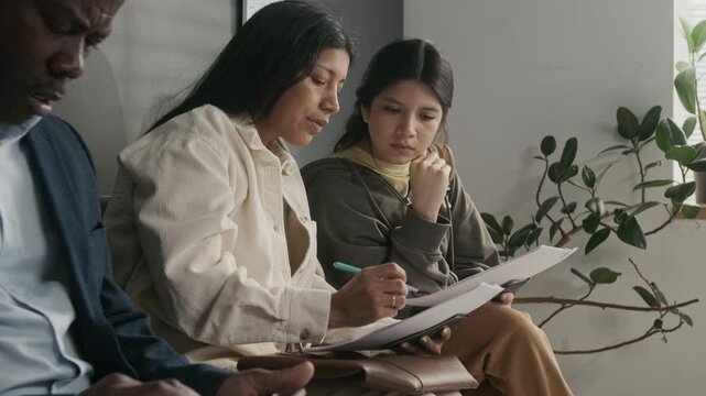 Side medium shot of Hispanic mother and daughter sitting in queue while waiting for appointment at foreign consulate, filling travel or study visa application forms next to black businessman in suit