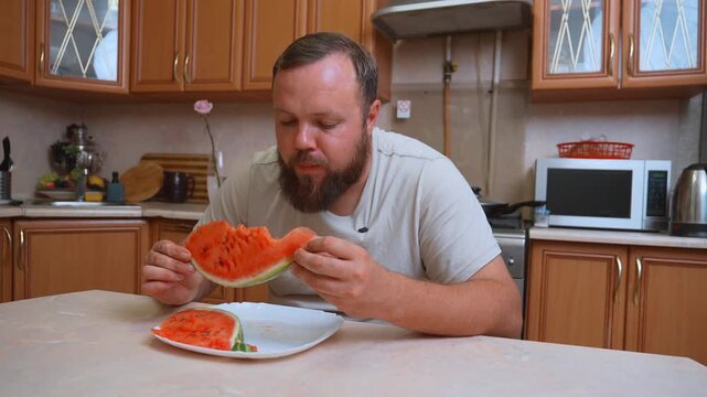 A hungry white chubby man with a beard sits at the table in the kitchen and greedily eats a large piece of ripe red watermelon, enjoying fresh and juicy summer fruits as a healthy snack