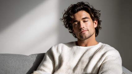 Obraz premium Serene young man with curly hair relaxing on a sofa with eyes closed, wearing a white knit sweater and holding a book in a sunlit minimalist home