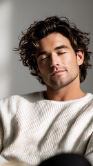 Fototapeta premium Serene young man with curly hair relaxing on a sofa with eyes closed, wearing a white knit sweater and holding a book in a sunlit minimalist home