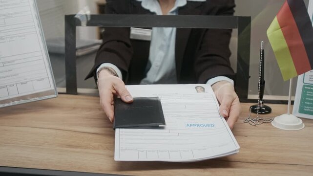 Midsection shot of unrecognizable female German consulate agent in black suit jacket handing back passport and visa documents with approval stamp to customer after successful submission