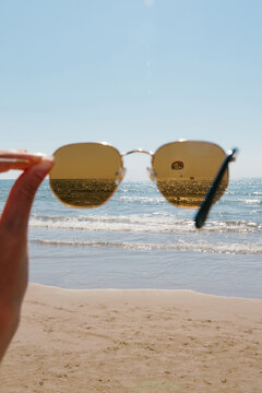 Hand holding sunglasses focusing on an ocean view, capturing a parasailer in the distance during a sunny day at the beach. Water sparkles under the clear blue sky. Summer travel creative concept.