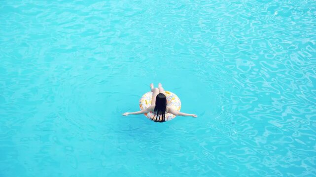 Young woman tourist in bikini swimsuit resting lies in rubber circle inflatable swimming lifebuoy ring floating on water in swimming pool, sunbathes thin beautiful body under summer sunlight, top view