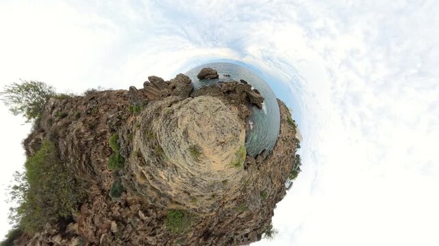 A rugged cliff juts into the sea, surrounded by water and steep rocks. The video is shot in a tiny planet effect, creating a spherical perspective of the coastal landscape.