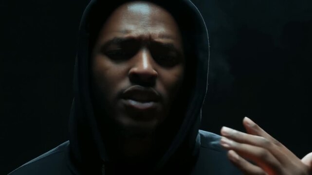Static close up shot of hooded man rapping and gesturing in dark smoke filled studio interior with dramatic low key lighting