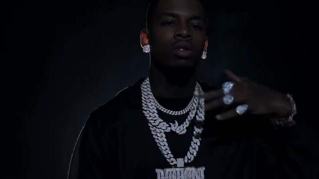Slow dolly in of man wearing layered chains gesturing with hand in dark studio with backlight spotlight