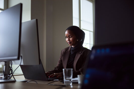 Female entrepreneur wearing headscarf working on laptop while sitting at desk in office
