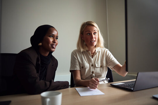 Smiling businesswoman sitting with female colleague explaining on computer at office desk