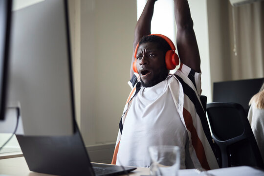 Tired male entrepreneur wearing wireless headphones yawning while stretching at desk in office