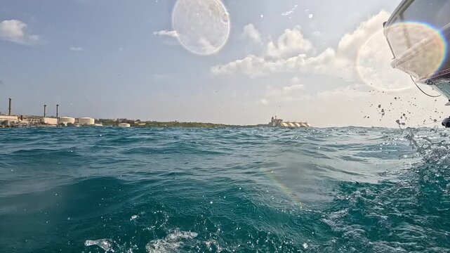 Low-angle shot from a moving boat showing turquoise sea splashes with the Delimara Power Station chimneys in the background in Malta