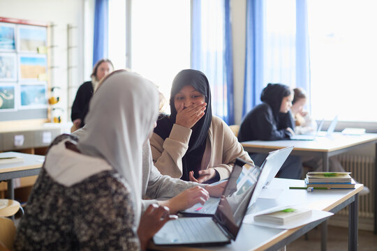 Girl in hijab covering mouth with hand while sitting at desk with friends in classroom
