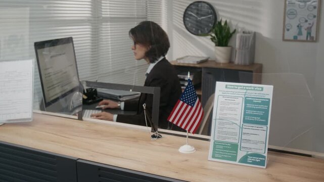 Medium side shot of Caucasian female USA visa agency officer sitting at desk behind glass partition counter, filling out application document on computer, American flag next to information leaflet