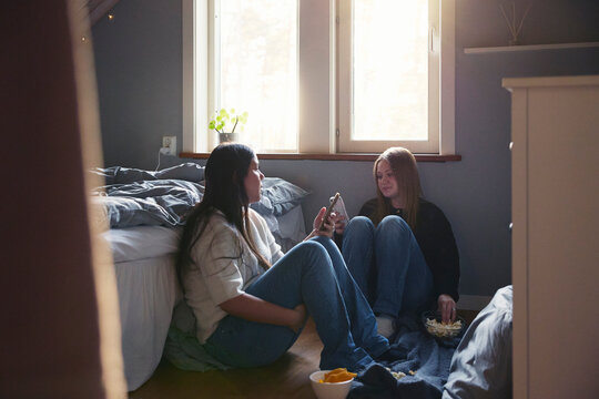 Teenage female friends using smart phones and having snacks while sitting in bedroom