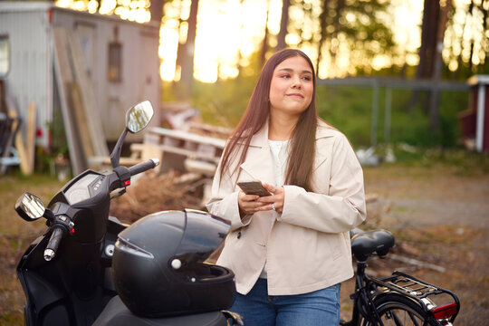 Smiling girl holding smart phone and looking away while standing by scooter