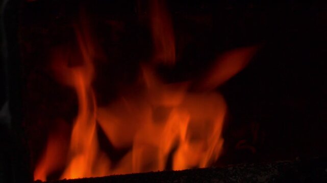 Close-up of bright flames burning inside a metal heating boiler. Dark background with subtle reflections of fire and a faint outline of the boiler door. Dramatic natural firelight and warm glow.