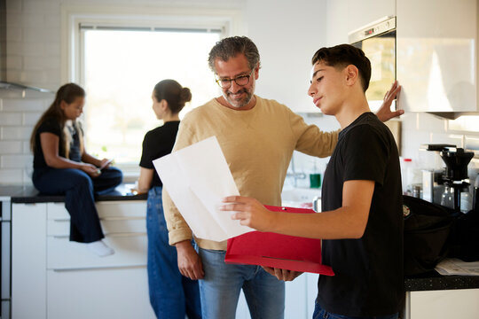 Smiling man looking at exam sheet of son while standing in kitchen