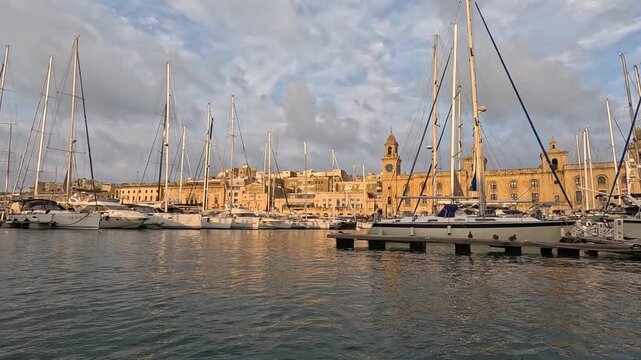Panning shot of luxury yachts moored at the Grand Harbour marina in Bormla (Cospicua) with the historic Malta Maritime Museum in the background during a golden sunset