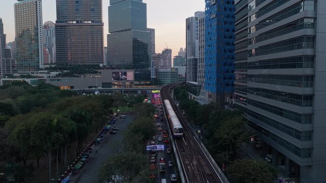 Aerial tracking view of BTS skytrain running on elevated tracks next to a busy street. Modern skyscrapers and green parks in Bangkok, Thailand. Bangkok, Thailand May 2. 2025