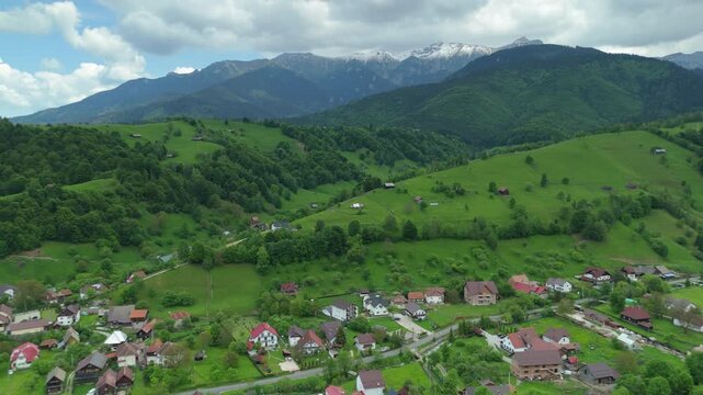 Picturesque rural village of Simon with modern houses and rustic wooden barns on vibrant green hills below the snow-capped Bucegi Mountains in Transylvania, aerial ascending shot