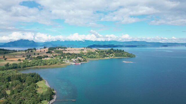 Panoramic pan over wide turquoise lake with forested and golden farmland peninsula revealing distant islands and Andean ranges under blue sky. Futrono, Chile