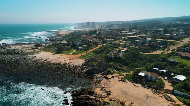 Drone shot over dramatic rocky headland with crashing waves and sandy coves revealing coastal residential town stretching along the Pacific shore. El Tabo, Chile